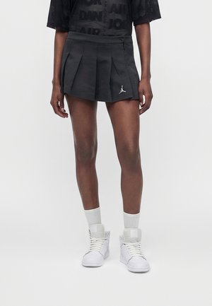 Osoba nosząca czarną plisowaną mini spódniczkę z logo Jordan, czarny graficzny t-shirt, białe skarpetki i białe sneakersy typu high-top na jednolitym tle.