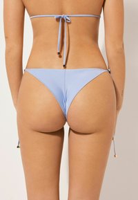 Blå bikiniunderdel med justerbara sidband, prydd med kvadratiska guldfärgade detaljer. Slät textur och minimal täckningsdesign.