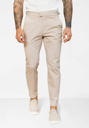 Beige tapered pants made of smooth fabric with a flat front, featuring side straps at the waist and subtle front pockets.