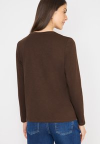 Braunes Langarmoberteil mit runder Ausschnitt, weicher Textur und lockerer Passform, kombiniert mit blauen Jeans, die am unteren Rand sichtbar sind.