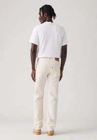 Hombre con cabello trenzado que lleva una camiseta blanca sencilla, jeans color blanco roto y zapatos marrones, de pie contra un fondo neutral, dando la espalda.