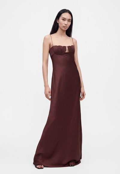 Femme grande aux longs cheveux foncés, portant une robe en satin marron sans manches avec de fines bretelles et des bonnets festonnés, debout devant un fond uni.