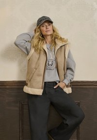 Beige schapenvachtvest met ritssluiting, grijze sweatshirt met print, zwarte wijde broek, en een grijze pet. Muur met subtiel patroon.