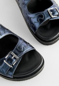 Blå metalflip-flops med udskæringer, sort sål og sølvfarvet spændehardware. Texturen er glat og skinnende.