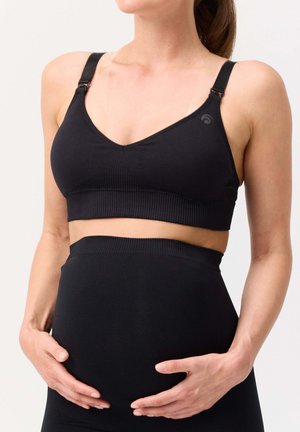 Schwangere Frau trägt schwarzen Umstands-BH und Leggings, ihre Hände halten sanft ihren Babybauch vor einem neutralen Hintergrund.