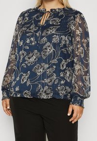 Blusa blu navy con maniche lunghe trasparenti, stampa floreale argento, scollatura arr gathered, e polsini elasticizzati. Vestibilità comoda con dettaglio a occhiello.