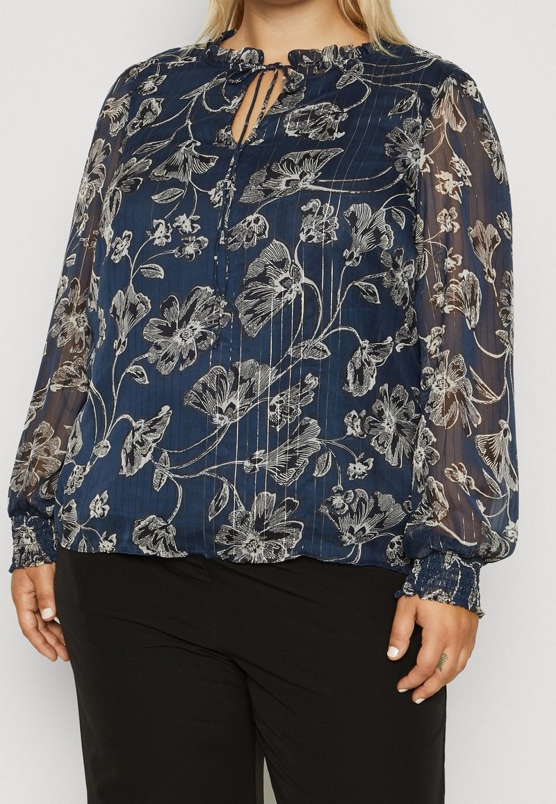 Blusa blu navy con maniche lunghe trasparenti, stampa floreale argento, scollatura arr gathered, e polsini elasticizzati. Vestibilità comoda con dettaglio a occhiello.
