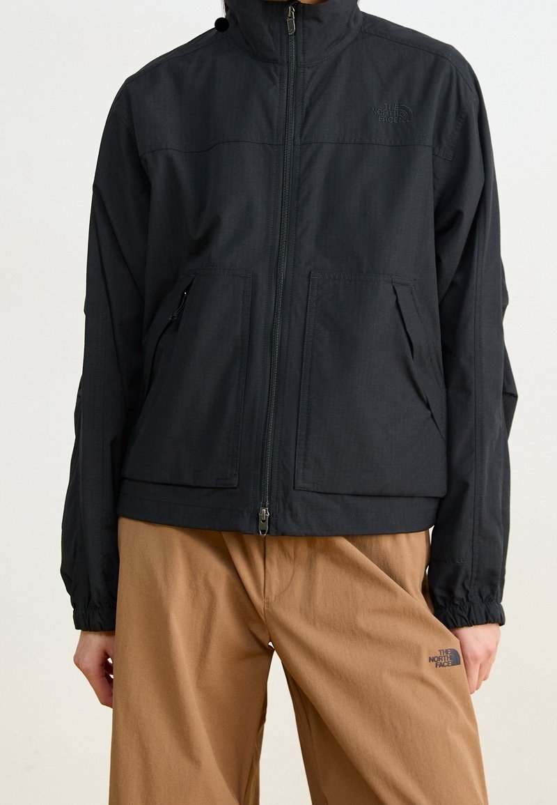 Svart zip-jacka med hög krage, tillverkad av textil med struktur. Har två framfickor och en logo på bröstet.