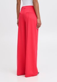 Pantalons à jambes larges en tissu rose vif, avec une texture lisse, des poches arrière et des détails de taille cintrée.