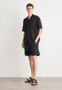 Chemise noire à manches courtes avec un motif en maille, associée à un short cargo noir doté d'une poche zippée sur le côté. Des sandales noires complètent le look.
