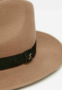 Beige felt hat with a wide brim, featuring a dark brown leather band and a decorative brass accent on the side. Soft texture throughout.