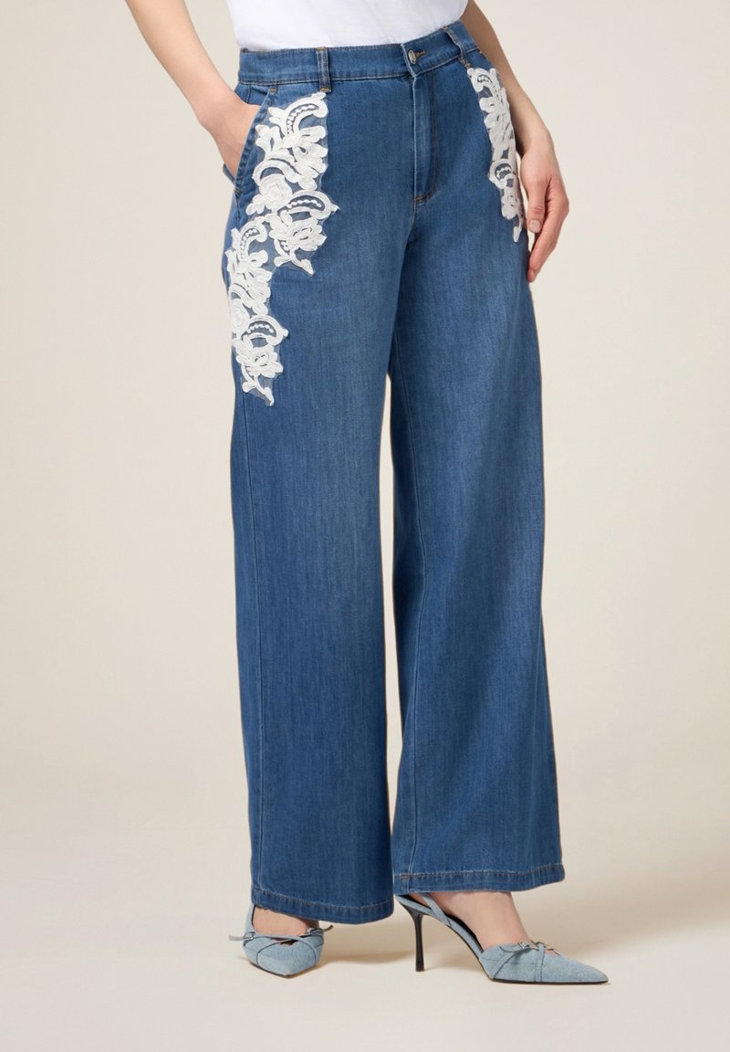 Jeans de mezclilla de pierna ancha en azul medio con bordado de flores de encaje blanco en los bolsillos delanteros. Presenta una cintura alta y diseño clásico de cinco bolsillos.