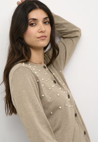 Beidzs kardigans, izgatavots no adīta materiāla, ar pērļu līdzīga dekora elementiem un tumšām pogām, brīvā fitā un garām piedurknēm.