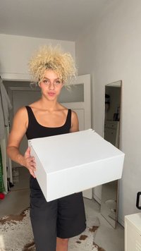 Hvid papkasse holdt foran en person, med et enkelt design, skarpe kanter og en mat finish. Baggrunden inkluderer et spejl og en dør.