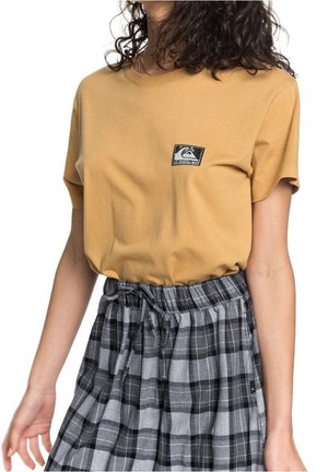 Kobieta nosząca musztardowożółty T-shirt z krótkim rękawem z małą czarno-białą naszywką, w połączeniu z wysokim stanem czarno-szarą spódnicą w kratę.