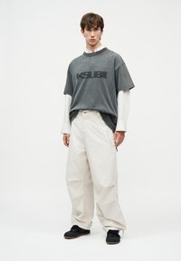 Jeune homme portant un T-shirt noir oversize déchiré sur une chemise blanche longue, un pantalon blanc large et des chaussures noires, debout devant un fond blanc uni.