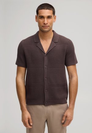 Junger Mann steht still, trägt ein kurzärmeliges, dunkelbraunes, strukturiertes Hemd mit Knöpfen und hellbeige Hose, neutraler grauer Hintergrund.