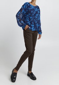 Blommönstrad blå blus med genomskinliga ärmar, kombinerad med bruna smala byxor och svarta loafers. Flera färger och släta texturer.