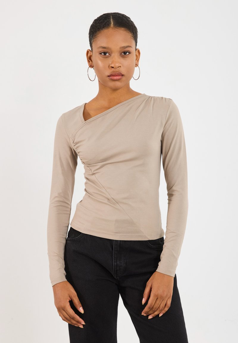 Maglietta beige a maniche lunghe con scollatura asimmetrica, dettaglio arricciato su un lato e tessuto morbido e liscio. Abbinata a pantaloni neri.
