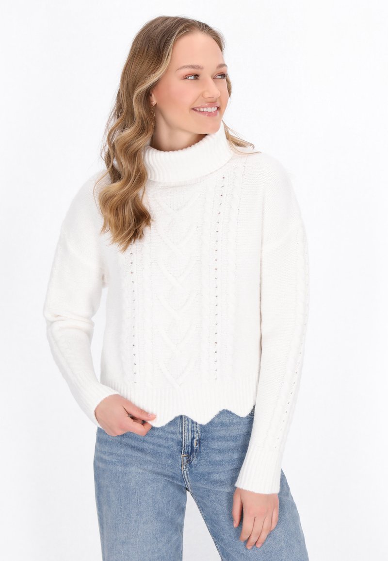 Weißer Kabelstrickpullover mit hohem, geripptem Kragen und gewelltem Saum, der ein strukturiertes Muster und lange Ärmel aufweist. Getragen mit blauen Jeans.