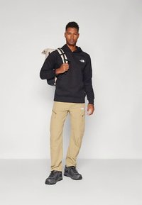 Man draagt een zwarte hoodie, beige cargobroek en wandelschoenen, met een rugzak over één schouder tegen een effen achtergrond.