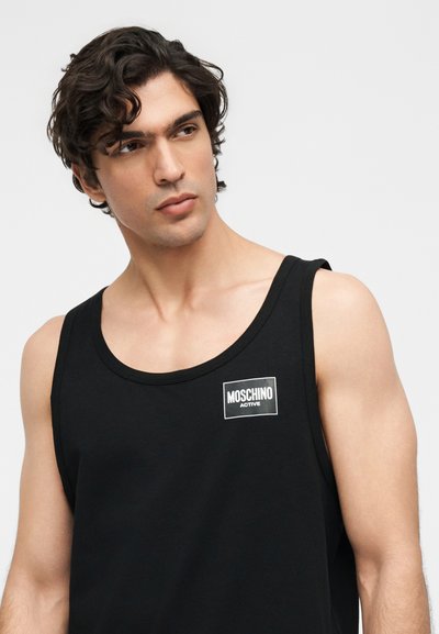 Άνδρας με κυματιστά σκούρα μαλλιά που φοράει μαύρο αμάνικο τοπ με λευκό λογότυπο "MOSCHINO ACTIVE" στο στήθος, κοιτάζοντας προς τα πλάγια.