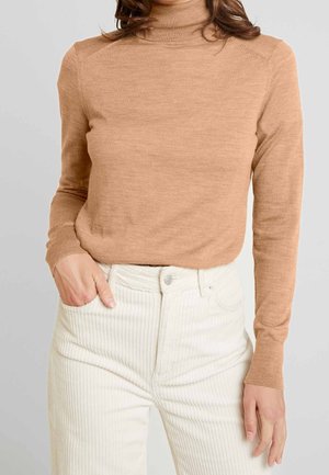 Donna con maglione a collo alto color cammello e pantaloni a vita alta in velluto a coste bianchi, con una mano in tasca, su sfondo grigio.