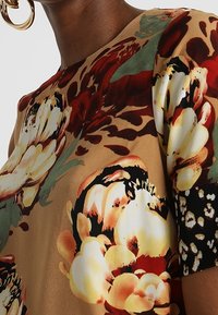 Blusa de estampado floral en beige, con grandes flores rojas y amarillas. Manga corta con acentos de estampado animal en blanco y negro. Textura suave.