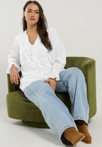 White, long-sleeve blouse with ruffle details, light blue denim jeans, and tan suede ankle boots, seated on a green upholstered chair.