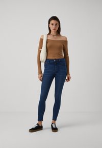 Top canelado castanho sem ombros, jeans skinny azuis e sapatilhas pretas com atacadores brancos. Leva uma bolsa de ombro de cor creme.
