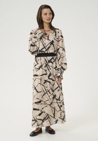Lange, geplooide jurk in lichtbeige met zwarte abstracte patronen. Heeft een cinched zwarte taille en uitlopende mouwen met delicate accenten.