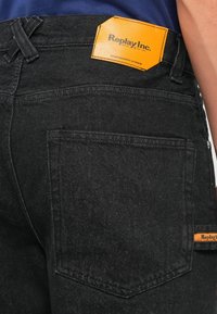 Vaqueros de mezclilla negra con una vista en primer plano del bolsillo trasero. Presenta una etiqueta de cuero naranja con el texto "Replay Inc." y costuras cuidadas.