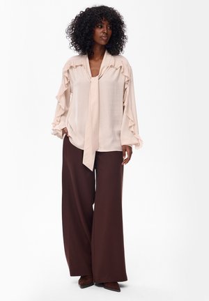Kobieta o kręconych włosach, ubrana w kremową bluzkę z falbankami i wiązaniem na szyi oraz szerokie brązowe spodnie, stoi z rękami w kieszeniach.