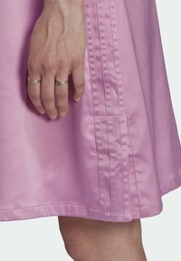 Sukienka z gładkiego, różowego satyny ma sylwetkę w kształcie litery A, subtelny połysk oraz pionowe zaszewki po bokach, co podkreśla jej elegancję.