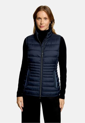 Vrouw draagt een marineblauwe doorgestikte mouwloze vest over een zwarte top met lange mouwen, staand voor een effen lichte achtergrond.