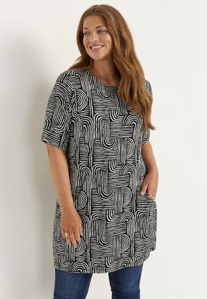 Kvinde med langt brunt hår, der smiler, iført en sort-hvid mønstret kortærmet tunika og blå jeans, stående mod en ensfarvet baggrund.