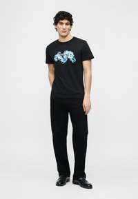 Άνδρας με σγουρά μαλλιά που φοράει μαύρο γραφικό T-shirt με μπλε σχέδιο, μαύρο παντελόνι και μαύρα δερμάτινα παπούτσια, που στέκεται μπροστά σε απλό φόντο.