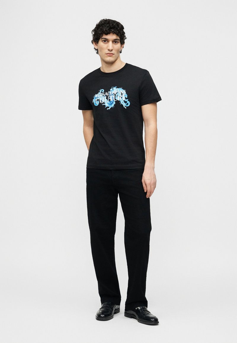 Άνδρας με σγουρά μαλλιά που φοράει μαύρο γραφικό T-shirt με μπλε σχέδιο, μαύρο παντελόνι και μαύρα δερμάτινα παπούτσια, που στέκεται μπροστά σε απλό φόντο.