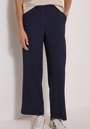 Femme portant un pantalon large bleu marine texturé et des baskets blanches, debout avec une main dans la poche sur un fond uni.