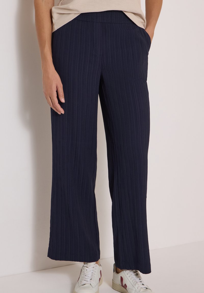 Femme portant un pantalon large bleu marine texturé et des baskets blanches, debout avec une main dans la poche sur un fond uni.