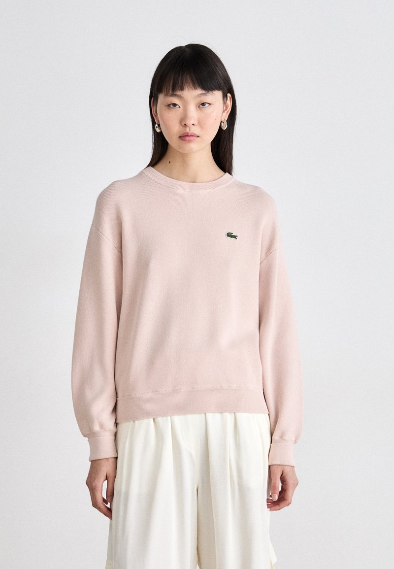 Suéter cor-de-rosa claro com decote redondo, punhos e barra canelados. Apresenta um pequeno logotipo verde da Lacoste no peito. O material parece suave.