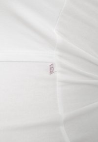 Valkoinen kangas, jossa näkyvä sauma ja pieni valkoinen etiketti, jossa on purppuranvärinen pieni "b"-logo ommeltuna lähelle reunaa.