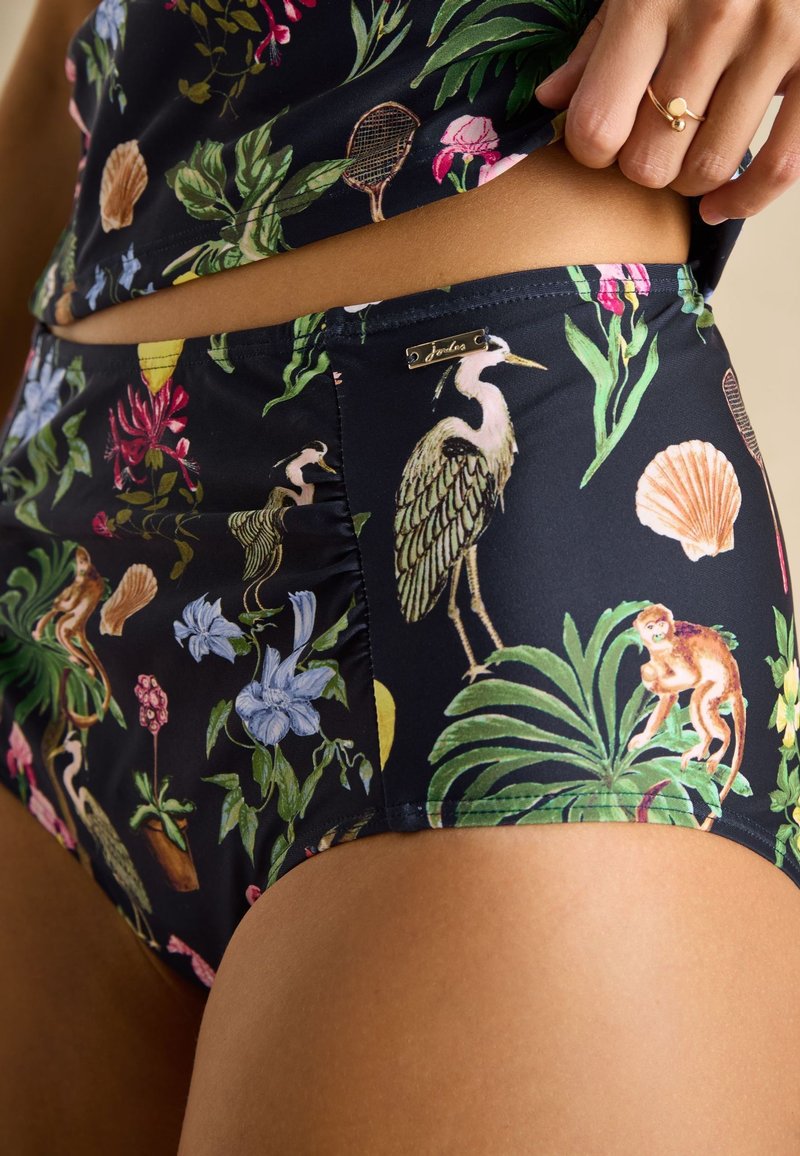 Nahaufnahme einer Person, die dunkle High-Waist-Hosen und ein Oberteil mit farbenfrohem tropischem Vogel-, Muschel-, Blumen- und Affenmuster trägt.