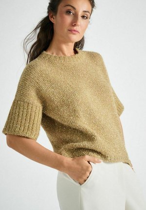 Kurzärmliger, goldfarbener Strickpullover mit geripptem Kragen und Bündchen. Hergestellt aus strukturiertem Stoff; leicht lockere Passform. Modell posiert mit der Hand in der Tasche.