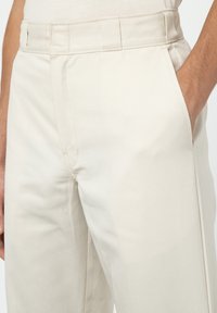 Detailaufnahme einer Person in hellbeigen Hosen, deren Hand teilweise in die rechte Tasche gesteckt ist, vor einem weißen Hintergrund.