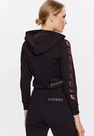 Donna con capelli biondi raccolti in uno chignon, che indossa una felpa nera con cappuccio e pantaloni con il testo "RICHMOND" in rosa e bianco sulle maniche e sul retro.