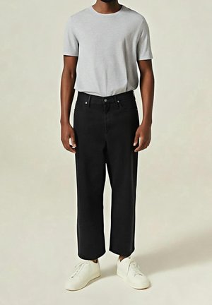Modèle masculin portant un t-shirt gris clair uni, un pantalon noir droit et des baskets blanches sur un fond neutre.