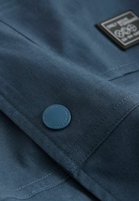 Sötétkék textil, rácsos mintázattal, kerek kék gombbal és fekete címkével, fehér szöveggel és olimpiai gyűrűkkel.