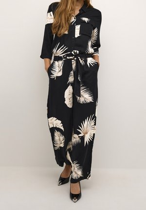 Musta, pitkähihaista haalarissa on valkoinen trooppisen lehden printti, vyö vyötäröllä ja rintatasku. Pintamateriaali vaikuttaa sileältä.