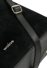 Zwarte handtas met een suède buitenkant, een gestructureerde leren schouderband en gouden hardware. Opvallend merkembleem in reliëf op de voorkant.